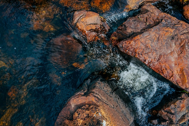 Glatte Wassernahaufnahme der Steine im Frühjahr. Sauberer Wasserfluss zwischen roten und orangefarbenen Steinen. Bunter natürlicher Gebirgsfrühlingsstrom mit copyspace. Schöne Beschaffenheit des Nebenflusses mit nassen Steinen.