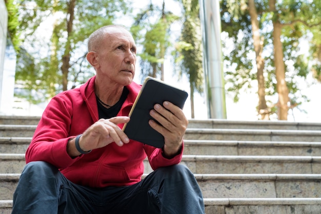 Glatzköpfiger älterer Mann, der digitales Tablett beim Sitzen auf der Treppe denkt und hält