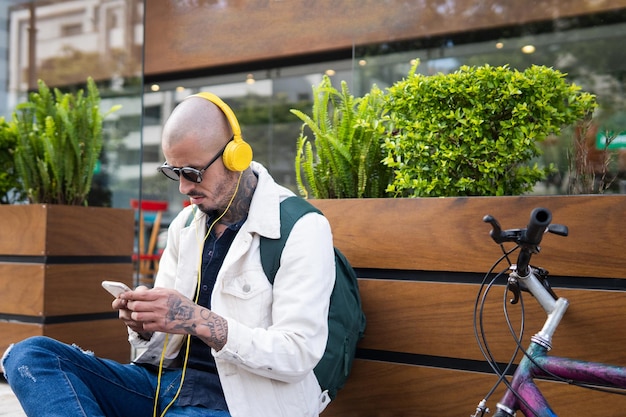 Glatzköpfiger Latino-Mann mit Tätowierungen, der mit Kopfhörern von seinem Handy aus Musik hört