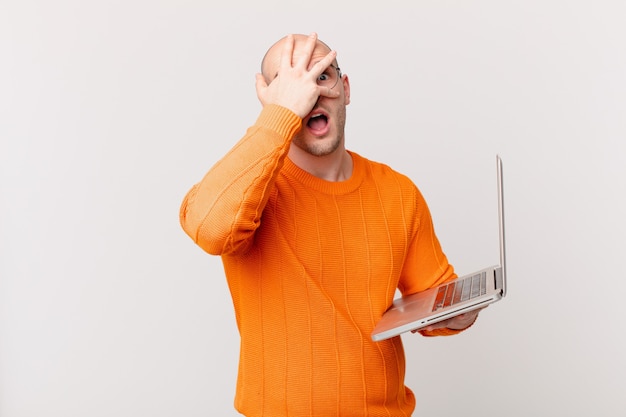 Glatzköpfiger Mann mit Computer, der schockiert, verängstigt oder verängstigt aussieht, das Gesicht mit der Hand bedeckt und zwischen den Fingern späht
