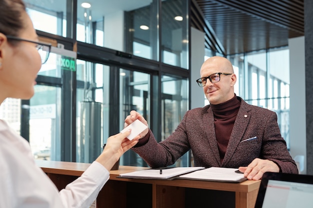 Glatzköpfiger reifer Geschäftsmann in Brille und Abendgarderobe, die Karte von der Hand der Empfangsdame nach dem Ausfüllen des Formulars nimmt