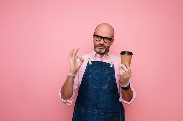 Glatzköpfiger reifer Mann, der OK-Symbol hält und Kaffee im recycelbaren Pappbecher hat