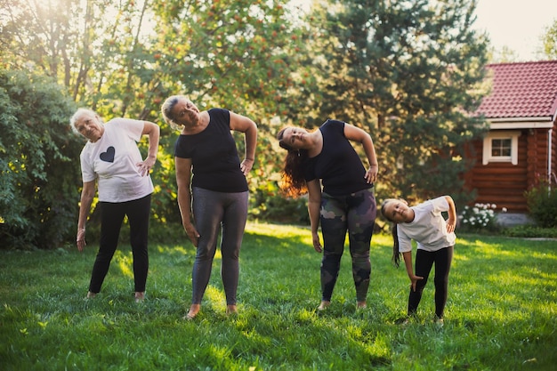 Gleiche Familie mit Frauen aus vier Generationen, die sich aufwärmen und sich strecken, um sich fit zu halten, auf einer Wiese voller grünem Gras und Bäumen Zeit miteinander verbringen