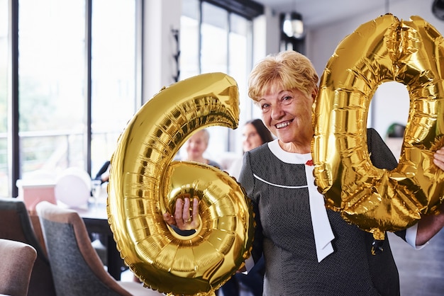 Con globos del numero 60 en manos. Mujer mayor con familiares y amigos celebrando un cumpleaños en el interior.