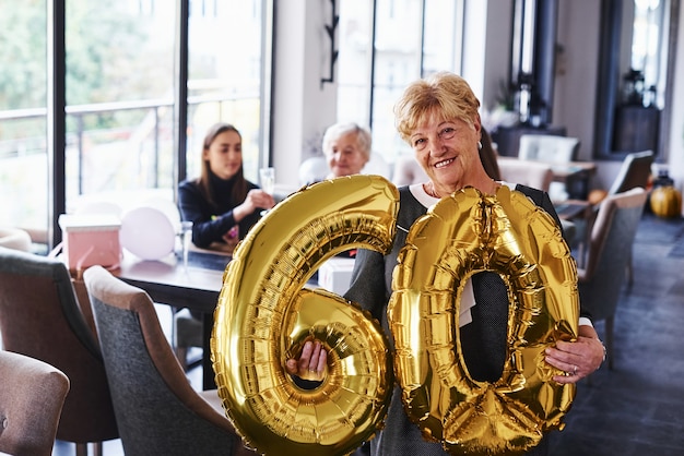 Con globos del numero 60 en manos. Mujer mayor con familiares y amigos celebrando un cumpleaños en el interior.