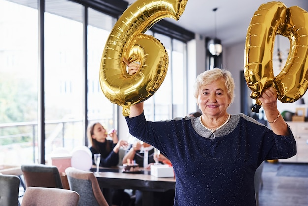 Con globos del numero 60 en manos. Mujer mayor con familiares y amigos celebrando un cumpleaños en el interior.