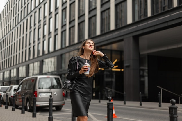 Glück elegante schöne Geschäftsfrau in Mode schwarzes Outfit mit Kleid und Ledertasche hält eine Tasse Kaffee und Spaziergänge in der Stadt in der Nähe eines modernen Gebäudes Städtisches lächelndes Mädchen