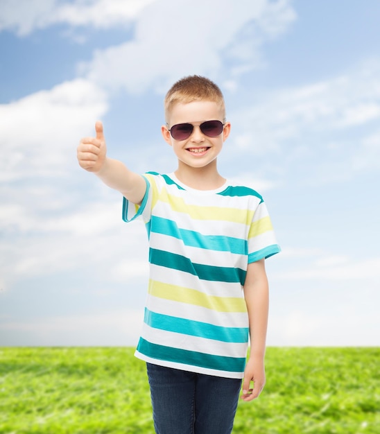 Glück, Sommer, Kindheit, Gesten und Personenkonzept - lächelnder süßer kleiner Junge mit Sonnenbrille über natürlichem Hintergrund, der Daumen nach oben zeigt