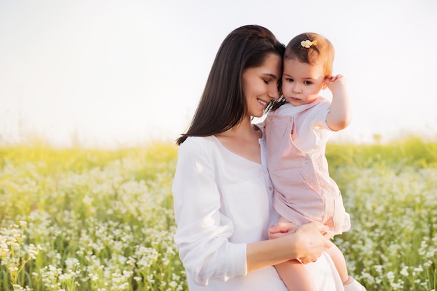 Glück und Zärtlichkeit von Mutterschaft und Kindheit Hübsche brünette Mutter umarmt eine Tochter Kleinkind Baby in den Händen lächelnd auf Blumenfeld Natur Hintergrund