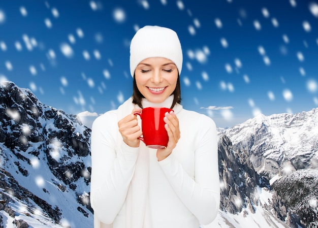 glück, winterferien, weihnachten, getränke und menschenkonzept - lächelnde junge frau in weißer warmer kleidung mit roter tasse über schneebedecktem berghintergrund
