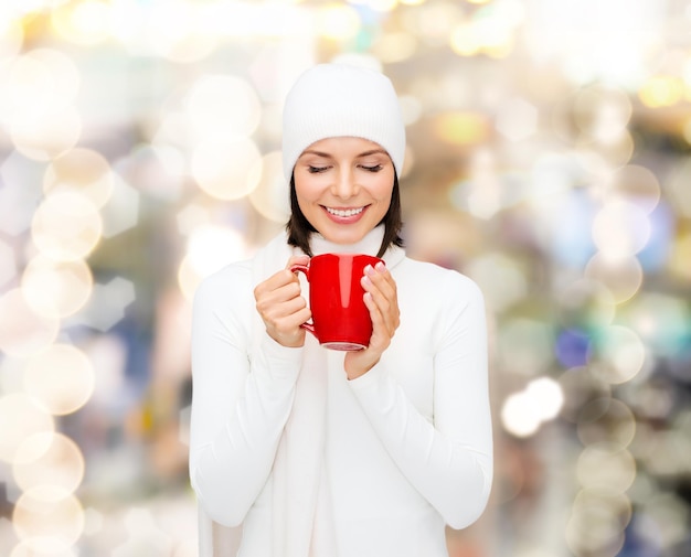 glück, winterferien, weihnachten, getränke und personenkonzept - lächelnde junge frau in weißer warmer kleidung mit roter tasse über lichthintergrund