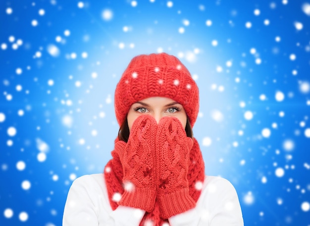 glück, winterferien, weihnachten und menschenkonzept - lächelnde junge frau in rotem hut, schal und handschuhen über blauem schneebedecktem hintergrund