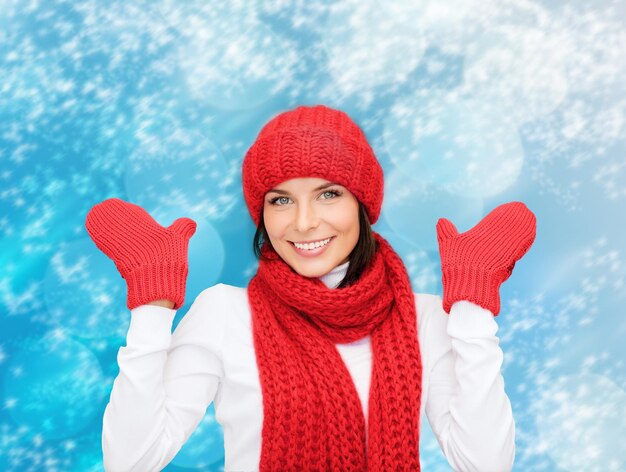 glück, winterferien, weihnachten und menschenkonzept - lächelnde junge frau in rotem hut, schal und handschuhen über blauem schneebedecktem hintergrund