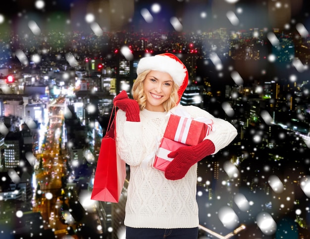 glück, winterferien, weihnachten und menschenkonzept - lächelnde junge frau in weihnachtsmannmütze mit geschenken und einkaufstasche über schneebedecktem nachtstadthintergrund