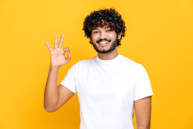 Glücklich angenehmer indischer oder arabischer lockerhaariger Typ in weißem T-Shirt, der ein Ok-Zeichen zeigt, während er auf einem isolierten orangefarbenen Hintergrund steht und freundlich lächelnd in die Kamera schaut