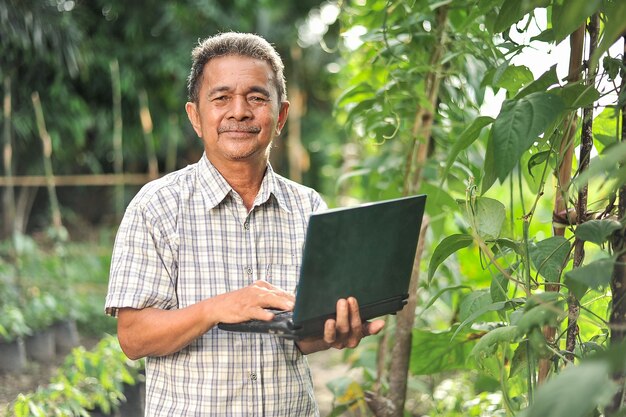 Glücklich des älteren asiatischen Bauernmannes, der Laptop auf dem Garten hält