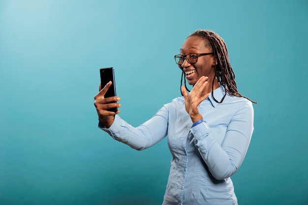 Glücklich lächelnde afrikanisch-amerikanische Frau mit Smartphone winkt bei Videokonferenz auf blauem Hintergrund. Schöne selbstbewusste junge Dame grüßt Freund über Online-Telefon-Videoanruf.