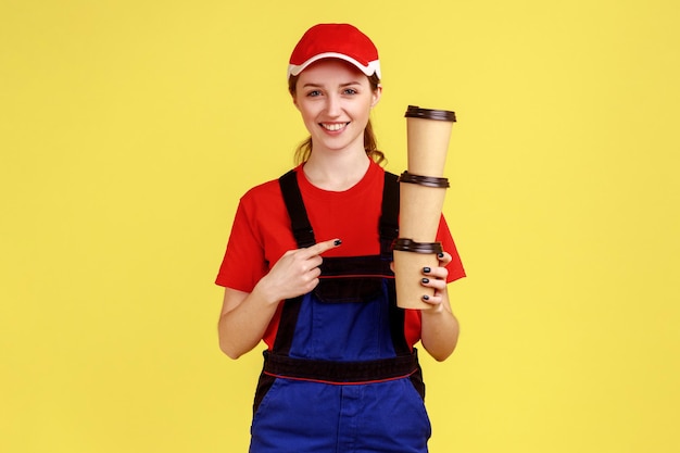 Glücklich lächelnde Arbeiterin, die auf einen Stapel von Einwegbechern Kaffee, Energiegetränk, Overall und roter Mütze hält und zeigt. Studioaufnahme im Innenbereich isoliert auf gelbem Hintergrund.