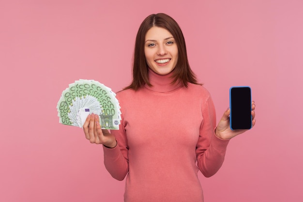 Glücklich lächelnde brünette Frau, die Spaß an Euro-Banknoten und Handy mit leerem Display in den Händen hält, mobile Banking-Anwendung. Studioaufnahme im Innenbereich isoliert auf rosa Hintergrund