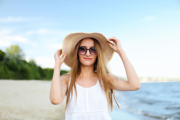 Glücklich lächelnde Frau in freier Glückseligkeit am Meeresstrand, die mit Hut und Sonnenbrille steht und posiert. Porträt eines weiblichen Models im weißen Sommerkleid, das während der Reise die Natur genießt