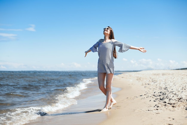 Glücklich lächelnde Frau in freier Glückseligkeit am Meeresstrand, die mit offenen Händen steht. Porträt eines brünetten weiblichen Models im Sommerkleid, das die Natur während der Reiseferien im Freien genießt, Gesamtplan.