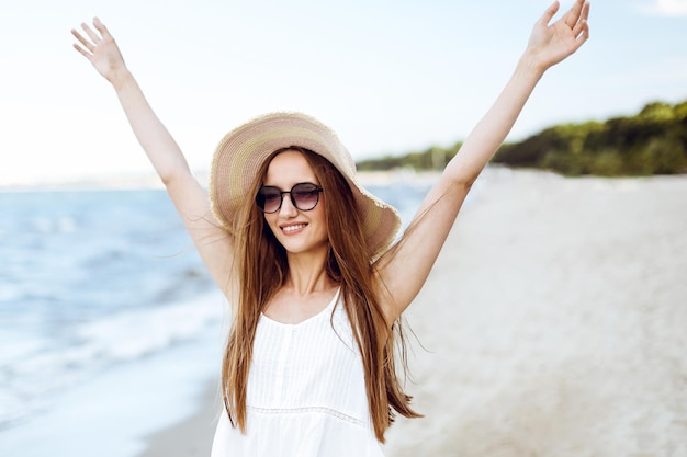 Glücklich lächelnde Frau in freier Glückseligkeit am Meeresstrand, stehend mit Hut, Sonnenbrille und erhobenen Händen. Porträt eines multikulturellen weiblichen Models im weißen Sommerkleid, das während einer Reise die Natur genießt