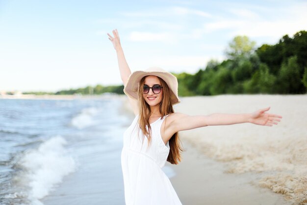 Glücklich lächelnde Frau in freier Glückseligkeit am Meeresstrand, stehend mit Hut, Sonnenbrille und erhobenen Händen. Porträt eines multikulturellen weiblichen Models im weißen Sommerkleid, das während einer Reise die Natur genießt