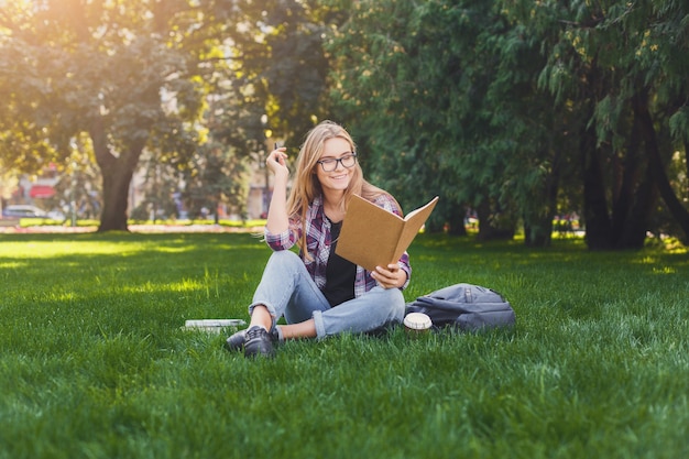 Glücklich lächelnde Frau liest Buch im Park, bereitet sich auf Prüfungen an der Universität oder Hochschule vor, sitzt auf Gras mit gekreuzten Beinen. Bildungskonzept, Kopienraum
