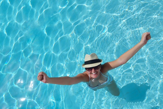 Glücklich lächelnde Frau mit Hut und Sonnenbrille im Swimmingpool im tropischen Resort