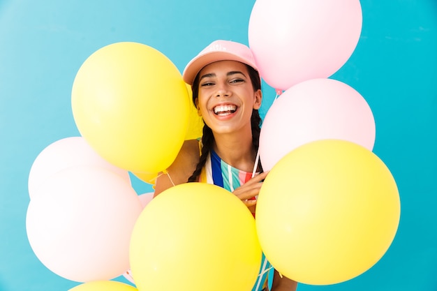 glücklich lächelnde Frau mit Mütze, die nach vorne schaut, während sie Luftballons isoliert auf blauer Wand hält?