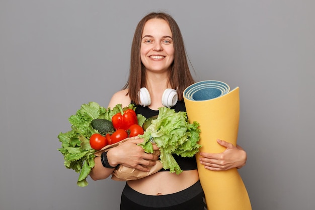 Glücklich lächelnde fröhliche junge Frau mit Gemüse und Yoga-Matte isoliert auf grauem Hintergrund umarmt Salat Tomaten Pfeffer gesunder Lebensstil