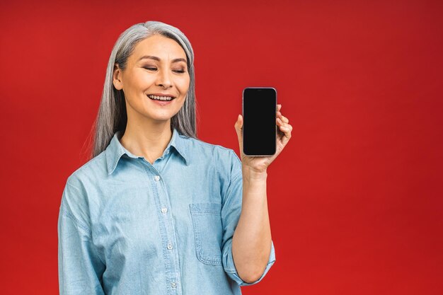 Glücklich lächelnde, glückliche asiatische Seniorin, reife Frau in lässigem, leerem Smartphone-Bildschirm, isoliert auf rotem Hintergrund Mit Telefon