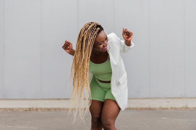 Glücklich lächelnde junge afroamerikanerfrau, die auf der straße tanzt. langes blondes geflochtenes haar. glück-konzept.