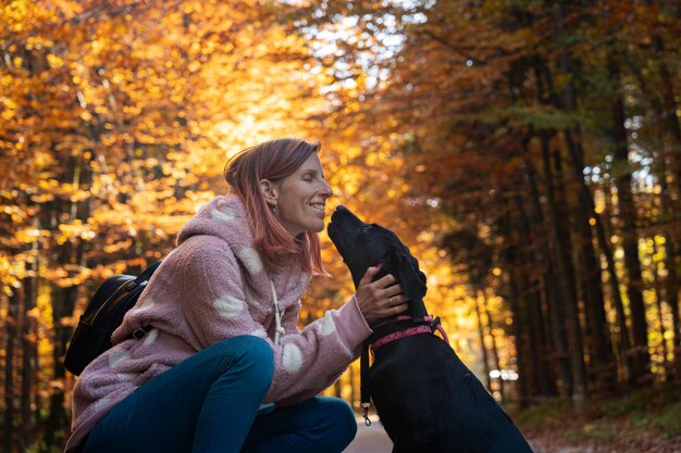 Glücklich lächelnde junge Frau, die sich hinkniet, um ihren süßen schwarzen Labrador-Welpen in der wunderschönen bunten Herbstnatur zu kuscheln, wobei Sonnenlicht durch die Bäume kommt.