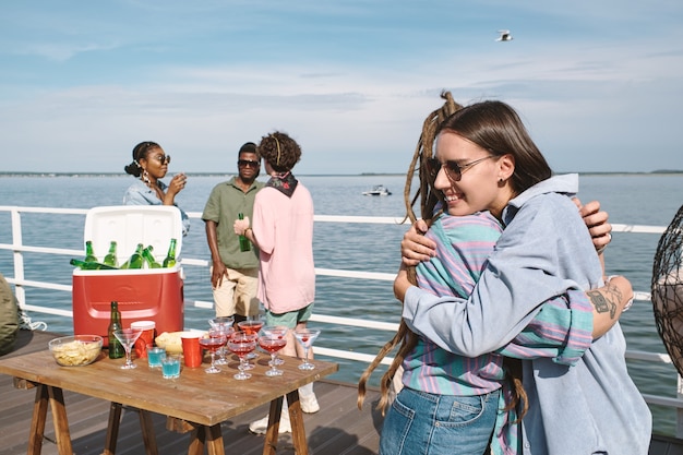 Glücklich lächelnde junge Frau mit Sonnenbrille, die ihre Freunde umarmt, die sie auf der Geburtstagsfeier am Pier getroffen hat?