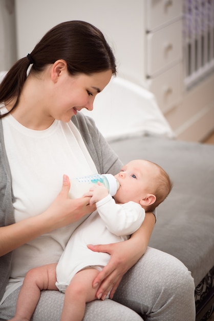 Glücklich lächelnde Mutter und Baby, die zu Hause auf dem Bett stehen und das Baby Milch isst.