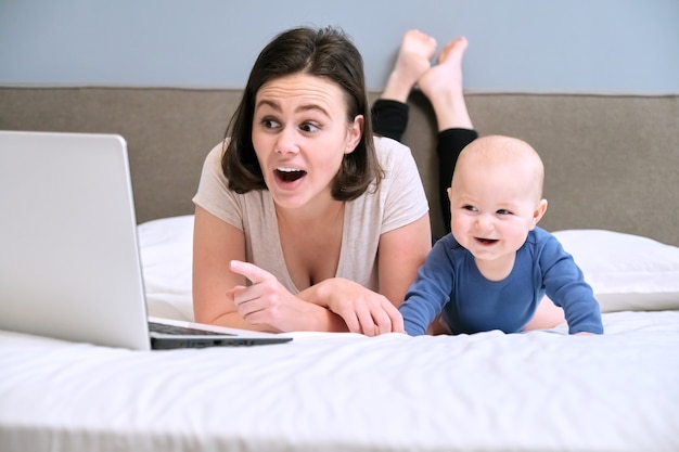 Glücklich lächelnde Mutter und Babysohn, die zu Hause zusammen auf dem Bett liegen und Laptop-Computer, Arbeit und Freizeit der jungen Mutter betrachten