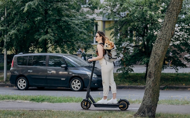 Glücklich lächelnde Reisende fährt mit ihrem Elektroroller in der Stadtparklandschaft mit dem Hund Welsh Corgi Pembroke in einem speziellen Rucksack