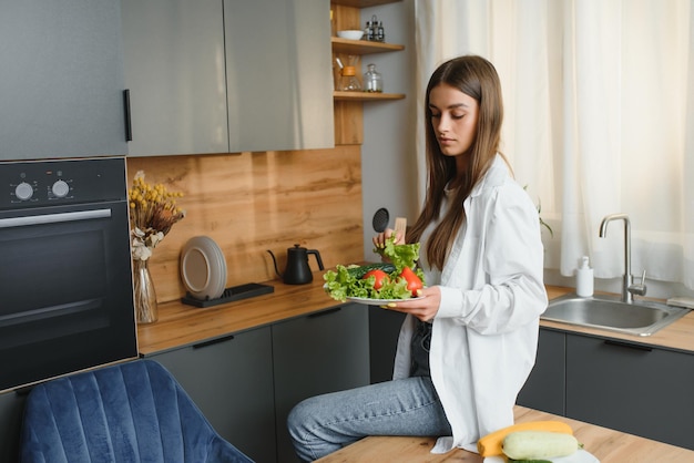 Glücklich lächelnde süße Frau bereitet einen frischen gesunden veganen Salat mit viel Gemüse in der Küche zu Hause vor und versucht ein neues Rezept