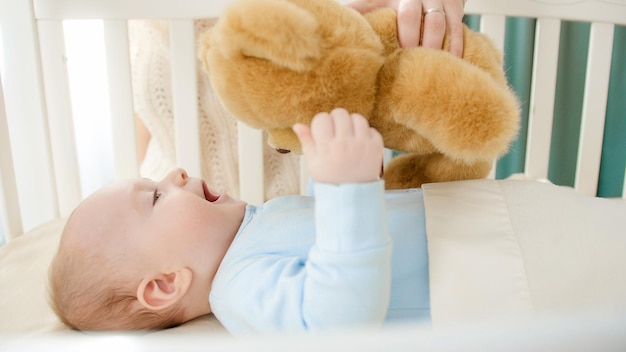 Foto glücklich lächelnder kleiner junge, der spielt und teddybär hält, während er in einer hölzernen wiege liegt. konzept der erziehung von familienglück und babyentwicklung