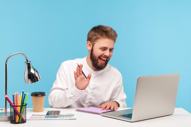 Glücklich lächelnder Mann Büroangestellter sitzt am Arbeitsplatz mit Notizblock und Bleistift und sagt hallo sprechender Videoanruf auf dem Laptop, der Online-Brainstorming-Meetings abhält Innenaufnahme isoliert auf blauem Hintergrund
