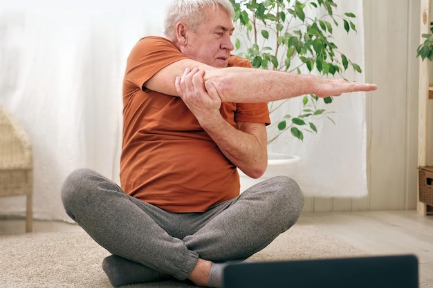Glücklich lächelnder reifer älterer Mann, der zu Hause Gymnastikübungen macht. Konzept eines gesunden Lebensstils, Fitness, Erholung, Wohlbefinden. Älterer Mann trainiert, Dehnübungen. Alter Mann trainiert