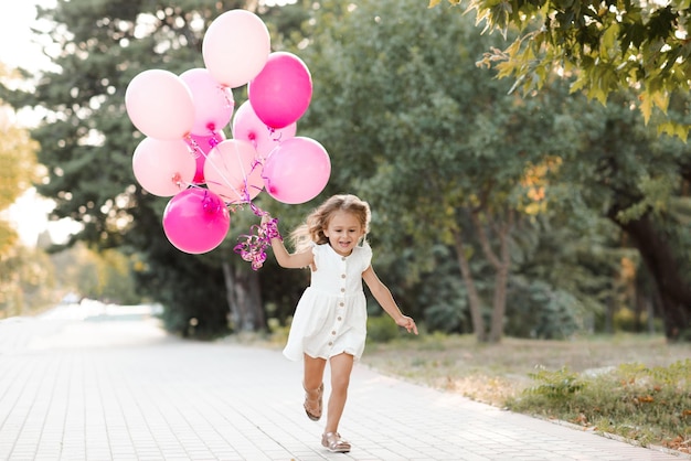 Glücklich lächelndes Kind Mädchen 3-4 Jahre alt tragen lässiges weißes Kleid mit rosa Luftballons laufen fröhlich