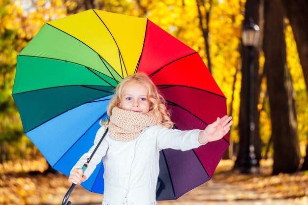 Glücklich lustig schöne Blondine mit blauen Augen Kind mit Regenbogen bunten Regenschirm im Herbst Park Mädchen Kind spielt auf der Natur im Freien Familienspaziergang im September Wald gelbe Bäume Blätter