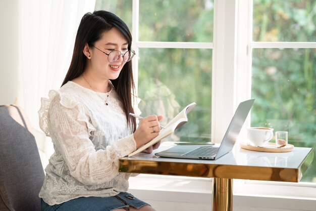 Glücklich von asiatischen freiberuflichen Menschen Geschäftsfrau, die schriftliche Notizen zum Notebook macht, beiläufig mit Laptop-Computer mit einer Kaffeetasse und Smartphone im Café arbeiten, Business Lifestyle