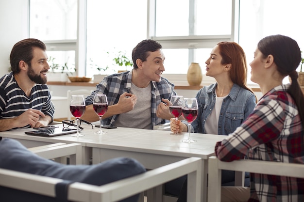 Glücklich zusammen. Gut aussehende inspirierte junge Freunde, die zusammen am Tisch sitzen, Wein trinken und reden