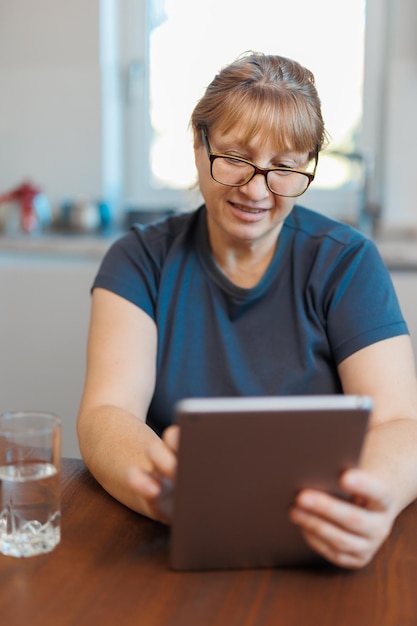 Glückliche 50-jährige Frau mittleren Alters mit digitalem Tablet, die zu Hause in der Küche sitzt