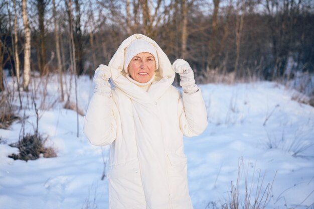 Glückliche ältere ältere reife Frau in der weißen warmen Oberbekleidung, die mit Schnee im sonnigen Winter draußen spielt