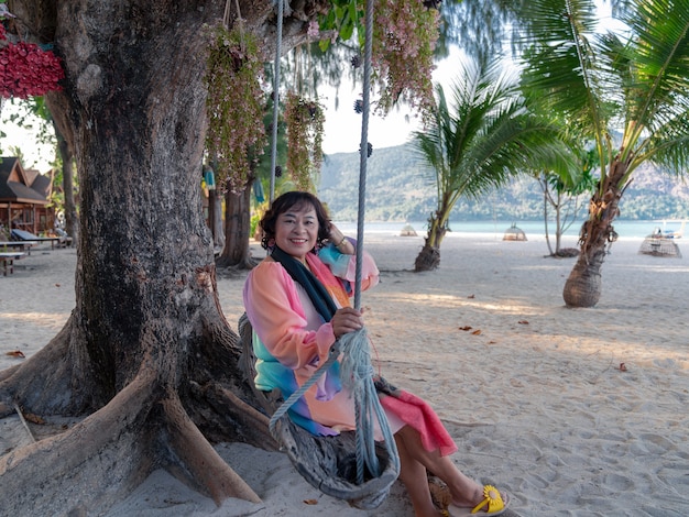 glückliche ältere asiatische frau in buntem kleid sitzt und entspannt sich auf der schaukel unter einem großen baum am strand