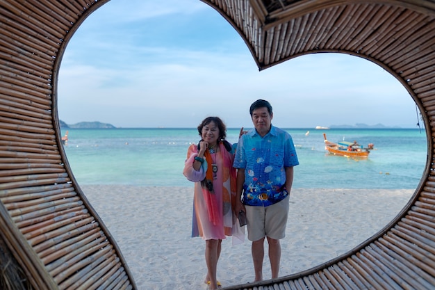 glückliche ältere asiatische Paarliebhaber, die am Strand hinter hölzernem Herzrahmen romantischen Lebensstil stehen